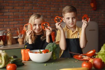Two children holding capsicum slices while preparing vegetables in a cozy kitchen setting.