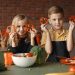 Two children holding capsicum slices while preparing vegetables in a cozy kitchen setting.