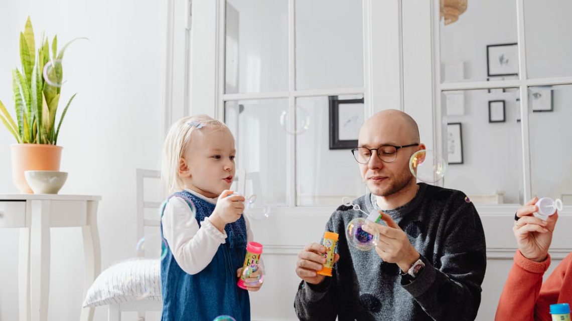 A joyful moment between a father and daughter blowing bubbles indoors, highlighting family fun.